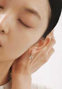 Pendientes de oro rosa con una gran piedra transparente facetada, rodeada de piedras más pequeñas, con un diseño simple y textura suave.