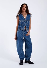 Kurteczka dżinsowa z głębokim dekoltem w kształcie litery V, zapinana na guziki, z dwoma przednimi kieszeniami, zestawiona z luźnymi spodniami dżinsowymi i czarnymi botkami za kostkę.