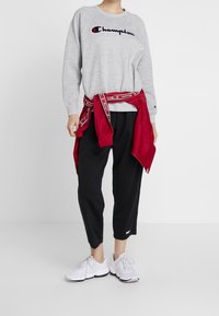 Felpa grigia con logo "Champion", giacca rossa legata in vita, pantaloni cropped neri e scarpe da ginnastica bianche con dettagli grigi.