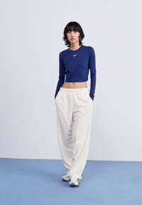 Hosszú ujjú, tengerészkék crop top, kis fehér logóval, világos krém színű, bő szárú melegítőnadrággal párosítva, amely rugalmas derékkal és oldalzsebekkel rendelkezik.
