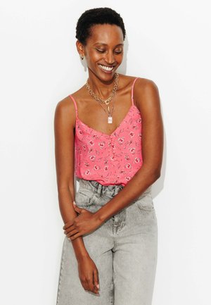 Femme souriante en caraco à fleurs rose et jean taille haute gris clair, portant des colliers en or superposés et des boucles d'oreilles créoles sur fond blanc.