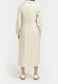 Pitkä beige mekko, jossa on kaulus, joustava vyötärö ja pitkät hihat. Pehmeä rakenne, joka laskeutuu kauniisti, käytetty valkoisten tennareiden kanssa.