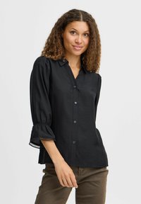 Camicia nera, leggera, con collo alla coreana, chiusura a bottoni e maniche a sbuffo, caratterizzata da un orlo arricciato e rifinita con pizzo.
