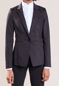 Blazer nero su misura con chiusura a un bottone, rever pronunciati e linee eleganti. Indossato sopra una camicia bianca con colletto, texture di tessuto liscia.