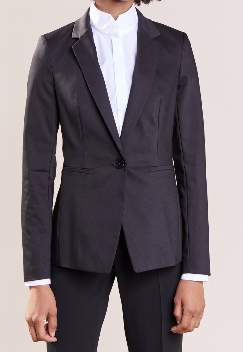 Blazer nero su misura con chiusura a un bottone, rever pronunciati e linee eleganti. Indossato sopra una camicia bianca con colletto, texture di tessuto liscia.