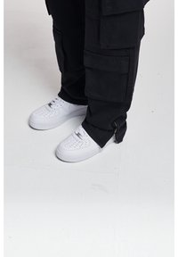 Baskets en cuir blanc avec des détails perforés, portées avec un pantalon cargo noir doté de multiples poches et d'une fermeture éclair à l'ourlet.