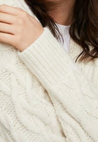 Woman wearing a textured cream-colored cable knit sweater, with her hand gently resting on her shoulder near dark brown hair.