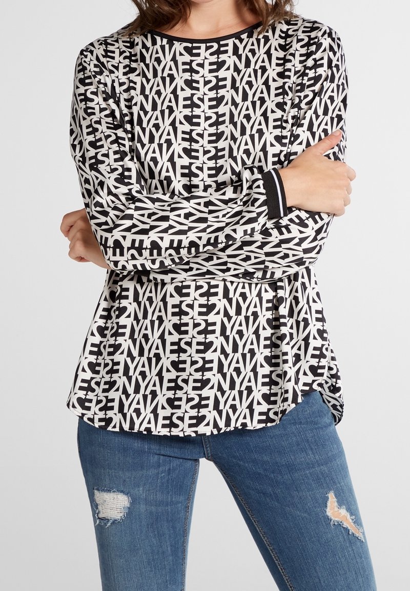Långärmad topp med ett svartvitt grafiskt textmönster, med rundad halsringning och kontrasterande svarta manschetter, i kombination med slitna blå jeans.