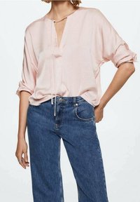 Blusa loose rosa chiaro con scollatura a V e dettaglio con fiocco, abbinata a jeans blu a vita alta. La blusa ha maniche arrotolate e una texture morbida.