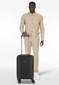 Maleta rígida negra con superficie texturizada y patrón, ruedas resistentes y asa retráctil. La lleva una persona con ropa de tonos neutros.