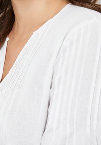 Femme portant un chemisier blanc avec des plis verticaux sur la poitrine et les manches, montrant l'encolure et le haut du torse.