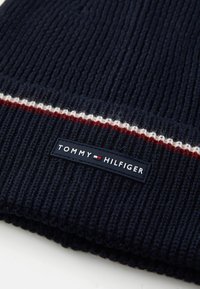 Mörkblå stickad mössa med ribbad textur, försedd med ett rödvitt stadband. Inkluderar en svart etikett med "TOMMY HILFIGER" text och logotyp.
