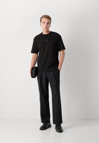 T-shirt noir à manches courtes avec logo, associé à un pantalon noir ample et des chaussures noires. La personne tient un petit sac noir.