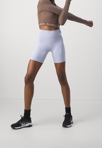 Frau, die ihre Arme ausstreckt und einen mauvefarbenen langärmeligen Cropped-Top, helllila hochtaillierte Radlerhosen, schwarze Socken und schwarze Nike-Sneaker trägt.