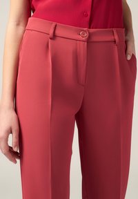 Pantaloni tailored rossi con texture liscia, caratterizzati da un'apertura frontale con bottone, tasche laterali e pieghe nette per una forma ben definita.