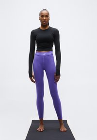 Αθλητική γυναίκα στέκεται ξυπόλυτη σε στρώμα γιόγκα φορώντας μαύρο crop top με μακριά μανίκια και μωβ κολάν σε ουδέτερη στάση.