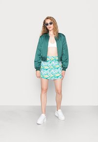 Casaco bomber verde, top cropped branco e uma saia curta com um padrão de ondas azuis e verdes. Ténis brancos completam o visual.