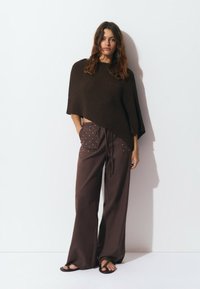 Poncho en tricot marron avec un ourlet asymétrique, associé à un pantalon marron ample avec des poches décoratives et une taille à cordon. Sandales noires.