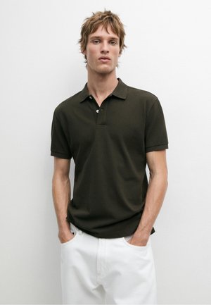 Jonge man met warrig haar, gekleed in een donkergroen poloshirt en witte broek, staat met handen in de zakken tegen een effen achtergrond.