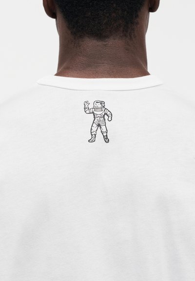 Λευκό βαμβακερό μπλουζάκι με μαύρο γραφικό αστροναύτη στο πάνω μέρος της πλάτης. Το σχέδιο περιλαμβάνει λεπτομέρειες όπως γάντια και φιμέ μάσκα.