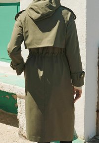 Oliwkowo-zielony płaszcz typu trencz z kapturem, skróconą górną częścią, elastycznym paskiem w talii oraz mankietami z guzikami. Gładka tkanina o minimalnej fakturze.