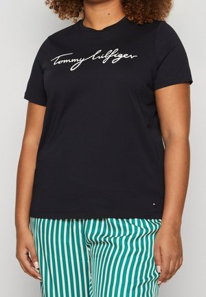 Person trägt ein schwarzes Tommy Hilfiger T-Shirt und grün-weiße vertikal gestreifte Hosen, steht vor einem einfarbigen Hintergrund.