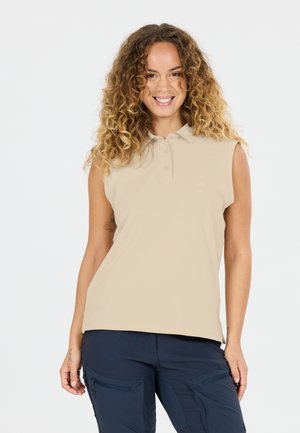 Smilende kvinde med krøllet hår iført beige ærmeløs poloshirt og marineblå cargobukser foran en ensfarvet hvid baggrund.