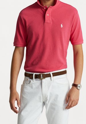 Man wearing coral pink polo shirt with white embroidered logo, white pants, brown belt, and wristwatch with brown leather strap.
