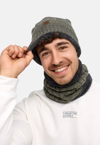 Knitted green beanie with fur lining and a logo patch, paired with a chunky ribbed neck gaiter in dark gray, worn by a smiling person.