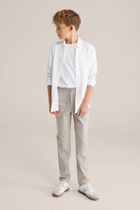 Witte button-up shirt over een witte t-shirt, gecombineerd met lichtgrijze jogger-achtige broek en witte sneakers met groene accenten.