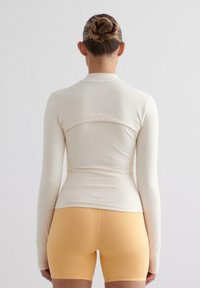 Kortermet kremfarget topp med en tettsittende design, som har en bakseam og høy krage, kombinert med gule shorts. Glatt tekstur, elastisk materiale.