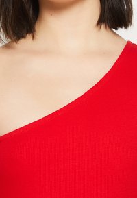 Haut rouge côtelé à une épaule avec un tissu texturé, présentant un large décolleté et une coupe ajustée, mettant en valeur l'épaule et la clavicule.