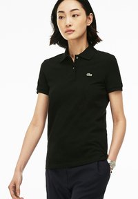 Svart polo skjorta med krage, korta ärmar och en grön Lacoste-logotyp på bröstet. Slät yta, kroppsnära design, enfärgad.