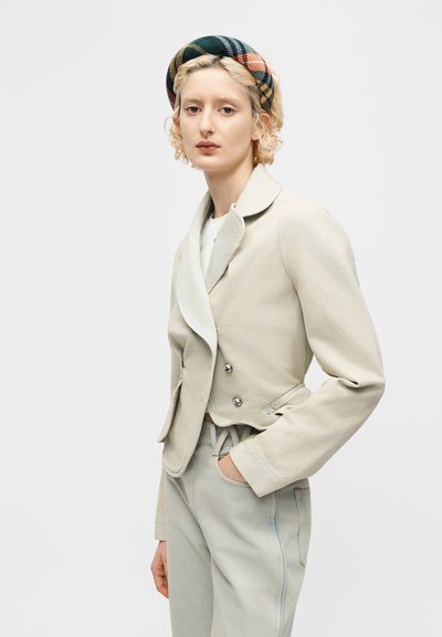 Mladá osoba s kučeravými blond vlasmi, oblečená v béžovom krátkom blejzri, svetlých nohaviciach a viacfarebnej kockovanej čelenke, stojaca s jednou rukou v vrecku.