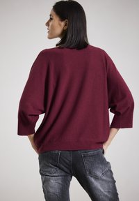 Burgunderfarbener Strickpullover mit lockerer Passform, überschnittenen Schultern und Dreiviertelärmeln, kombiniert mit dunklen Jeans.