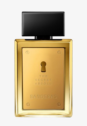 Flacon de parfum rectangulaire doré avec bouchon noir, forme de serrure à l'avant, et texte "The Secret Absolu Banderas."