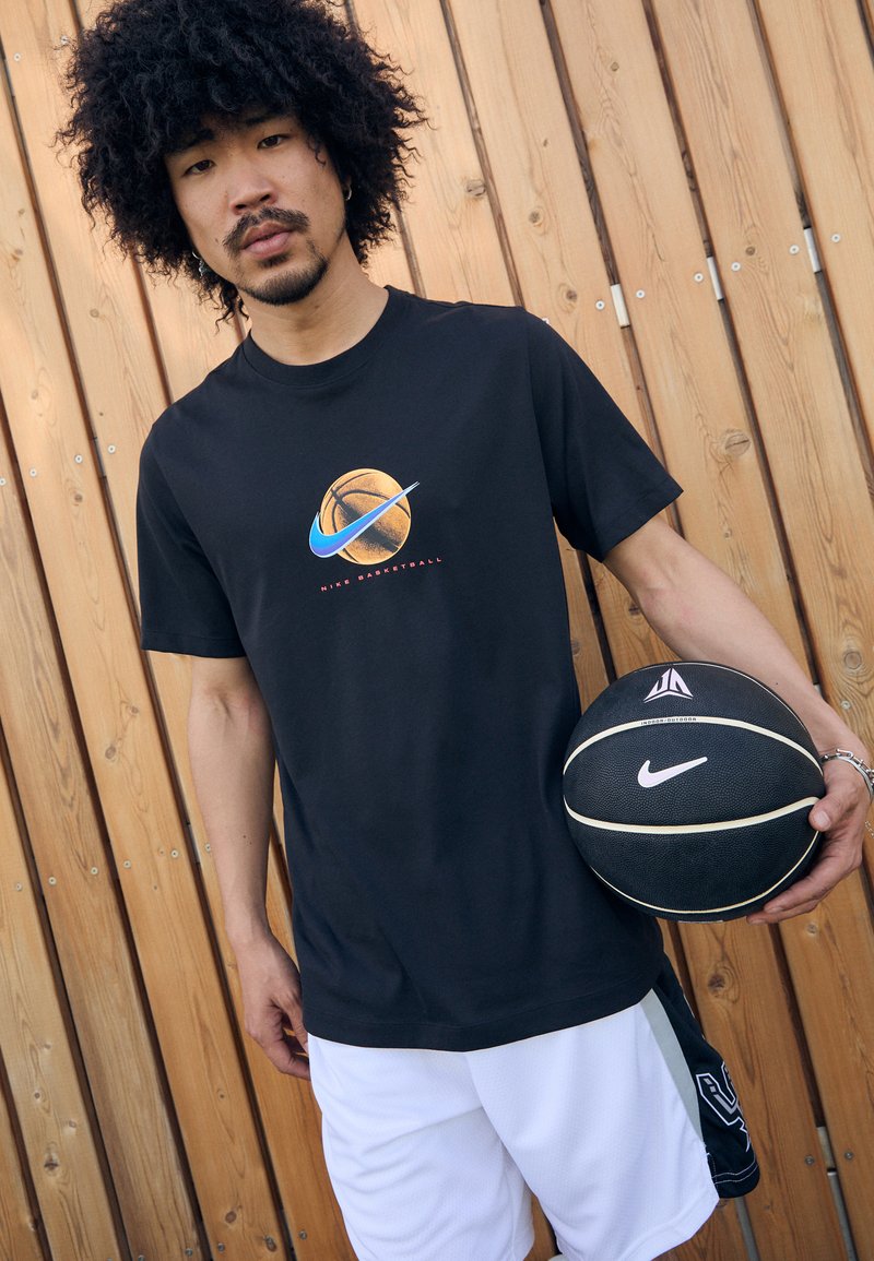 Camiseta negra de Nike con gráfico de baloncesto, pantalones cortos de baloncesto blancos, sosteniendo un baloncesto negro con acentos en blanco y gris, fondo de madera.
