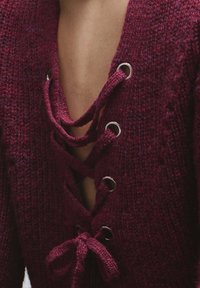 Jersey de punto color burdeos con un escote en V profundo, adornado con ojales metálicos y detalles de cordones, acentuado con un lazo en la cintura.