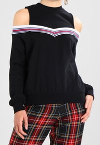 Svart kallskulder-topp med vit, röd och blå randdetalj, i kombination med röda och svarta rutiga byxor, händerna vilande vid sidorna.