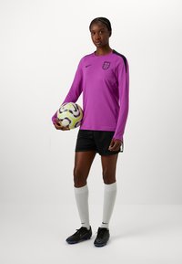Fialový dlouhý rukáv dres s černými akcenty, černé kraťasy, bílé vysoké ponožky a černé fotbalové kopačky, držící multicolor fotbal.