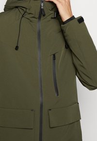 Giacca verde oliva con finitura opaca, caratterizzata da una chiusura a zip sul fronte, due grandi tasche a pattina e cordini regolabili su cappuccio e polsini.