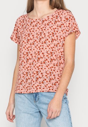 Femme portant un chemisier rose clair à manches courtes avec un petit imprimé floral rouge et un jean en denim bleu clair sur un fond uni.