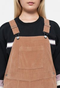 Salopette in corduroy di un morbido colore pesca tenue, con quattro bottoni, spalline regolabili e una tasca frontale. Indossata sopra una maglietta nera.