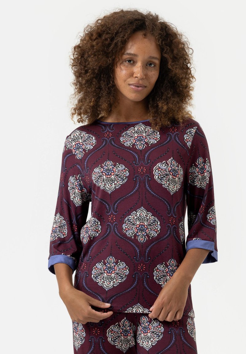 Bordeaux top met bloemig patroon in wit, blauw en rood. Heeft driekwartmouwen met blauwe manchetten en een ronde halslijn. Zachte textuur.