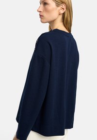 Granatowy sweter z długim rękawem, wykonany z miękkiego dzianiny, o luźnym kroju, z guzikami na jednym ramieniu, okrągłym dekoltem i ściągaczem u dołu.