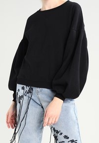 Sudadera negra con cuello redondo y mangas voluminosas, combinada con unos vaqueros de color azul claro que tienen texto impreso y detalles desflecados.