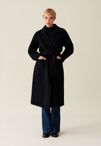 Cappotto nero a maniche lunghe con vita cintata, colletto a scollatura ad angolo e due tasche frontali; indossato sopra jeans svasati blu e scarpe nere.