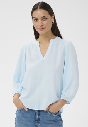 Žena s dlhými tmavými vlasmi má na sebe svetlomodrú blúzku s výstrihom do V a tromi štvrtinovými nafúknutými rukávmi a modré džínsy, ruky v kapse, mierne sa usmieva.