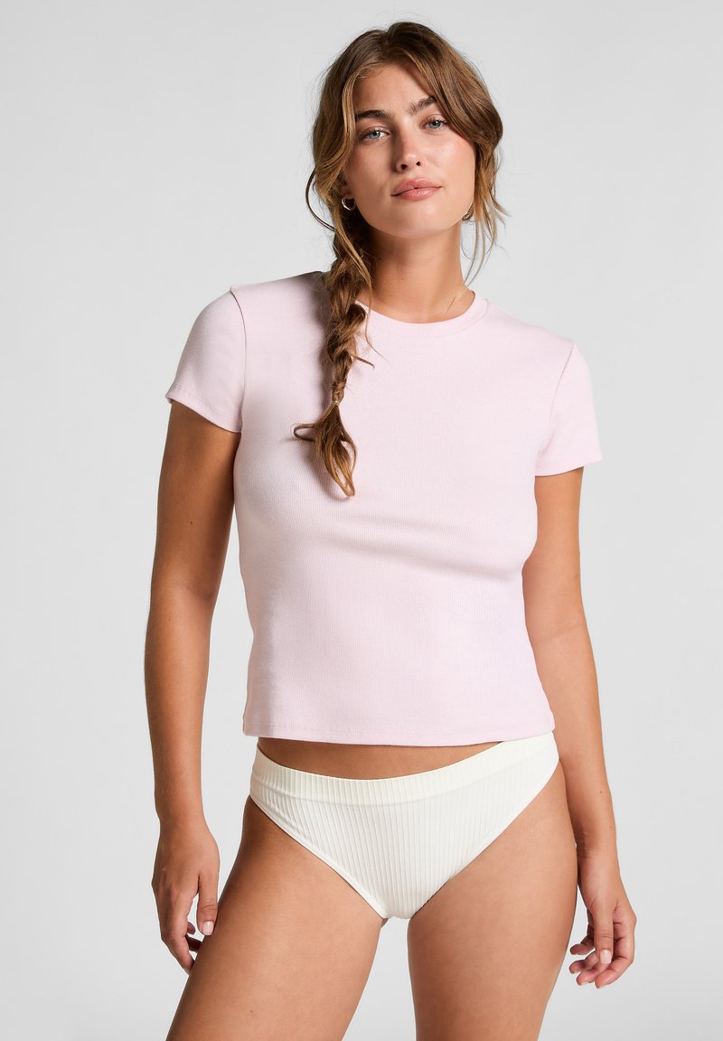Vrouw met gevlochten haar, gekleed in een lichtroze korte mouw crop top en hoog uitgesneden witte gerekte onderbroeken tegen een effen achtergrond.
