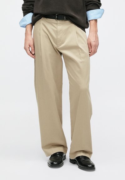 Šviesiai rudos plačios kojų kelnės su raukiniais, pagamintos iš lengvos medžiagos. Jos derinamos su tamsiu megztiniu ir juodais odiniais batais.